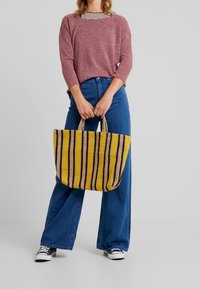 Grote geweven shopper met gele en zwarte strepen, gecombineerd met een losse roze trui en wijde blauwe jeans. Casual schoeisel erbij.