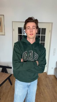 Ung mand stående indendørs med armene over kors, iført en mørkegrøn GAP-hoodie og lyseblå jeans, foran hvide dobbeltdøre.