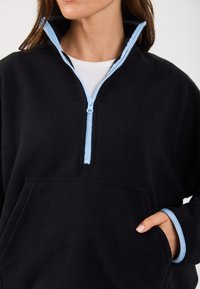 Pulover negru din fleece cu fermoar pe jumătate, accentuat cu albastru deschis. Dispune de un buzunar tip kangur și manșete ribbate, oferind o textură moale.