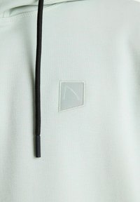 Sudadera verde claro con tejido texturizado, que presenta un pequeño parche con logo y un cordón negro. Textura suave, diseño geométrico sencillo.