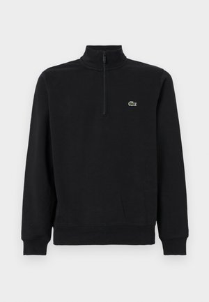 Svart fleecepullover med hög krage, halvdragkedja och en liten grön krokodil-logotyp på bröstet. Mjuk, strukturerad textur.