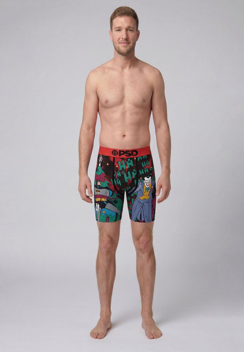 Junger Mann steht barfuß und trägt bunte Boxershorts mit Batman- und Joker-Motiv mit einem roten Bund mit der Aufschrift PSD vor einfachem Hintergrund.
