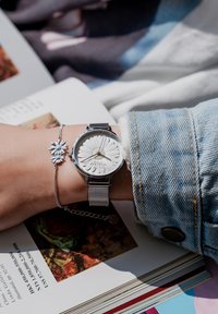 Orologio con cinturino in maglia argentata e quadrante floreale bianco testurizzato su un polso, abbinato a un delicato bracciale argentato, poggiato su un libro aperto.