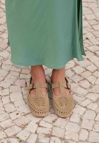 Sandalias de ante marrón con diseño trenzado, cortes abiertos y hebillas ajustables, combinadas con una falda verde fluida sobre un pavimento de piedra.