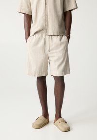 Beige linnen overhemd met knopen en een relaxed fit, gecombineerd met shorts met elastische taille in een vergelijkbare tint. Suède instappers met een gestructureerde afwerking.