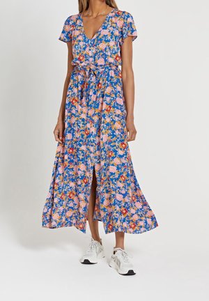 Femme portant une robe bleue à fleurs avec col en V, manches courtes, taille nouée, boutons sur le devant et baskets blanches sur un fond uni.