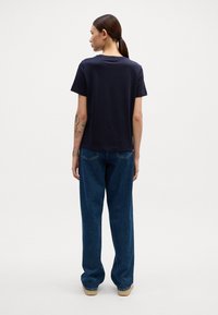 Kort marinblå T-shirt med rund halsringning, i kombination med lösa, blå denimjeans. Anmärkningsvärda tatueringar synliga på armen; enkel, avslappnad design.