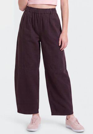 Vrouw draagt losse donkerpaarse hoge taillebroek met elastische tailleband en lichtroze sneakers op witte achtergrond.