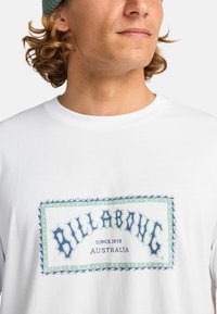 Hvid bomulds t-shirt med et stort, farverigt "Billabong" logo med blå og grønne accenter og en dekorativ kant.