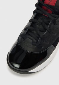 Zwarte sportschoen met een neuskap van lakleer, een gestructureerd bovenwerk, zwarte veters en rode accenten op de tong, met geperforeerde details.