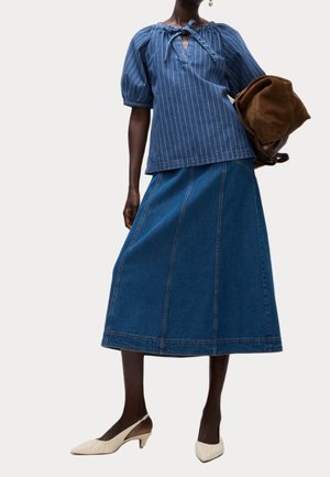 Vrouw draagt een blauw gestreepte top met pofmouwen, een blauwe denim midirok, crème slingback kitten heels, en houdt een opgevouwen bruine tas vast.