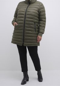 Oliivinvihreä toppatakki, jossa on korkea kaulus, vetoketjusuljenta ja kaksi sivutaskua. Takissa on vaakasuora tikkaus ja mustat vetoketjuaccentit.