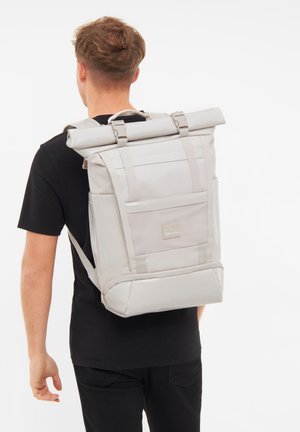 Zaino roll-top grigio chiaro con spallacci regolabili, tasche in rete e design rettangolare, caratterizzato da un logo sul pannello frontale.