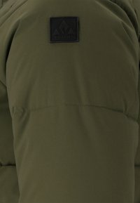 Olijfgroen geïsoleerde jas met een gestructureerd oppervlak, een gequilt patroon en een opvallende zwarte logo patch op het bovenste gedeelte.