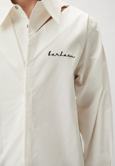 White long-sleeve shirt with a pointed collar, featuring a cut-out detail on one side and black embroidered text "berhasm" on the chest.