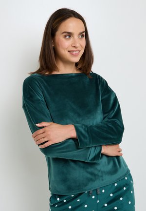 Maglia lunga in velluto color teal con scollo a barca, caratterizzata da una texture liscia. Indossata con pantaloni a fantasia di pois. Modella con le braccia incrociate.
