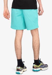 Shorts turquoise avec une texture lisse, présentant une coupe droite et des poches latérales, assortis à des chaussures de course multicolores et des chaussettes noires.