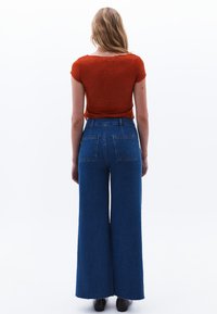 Červený krajkový top s krátkým rukávem spárovaný s modrými džínami s vysokým pasem a širokými nohavicemi. Hladká textura džínů se dvěma zadními kapsami.