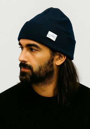 Navy knit beanie with ribbed texture, featuring a white logo patch. The cap is fitted, tapering at the top for a snug fit.
