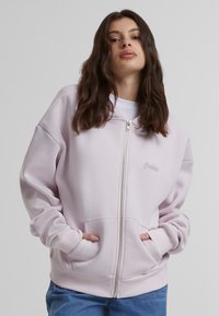 Felpa rosa con zip realizzata in tessuto morbido, dotata di tasche frontali, polsini e orlo a costine, con la parola "Proibito" ricamata in viola chiaro.