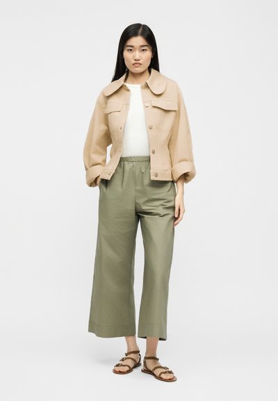 Γυναίκα που στέκεται φορώντας μπεζ cropped σακάκι, λευκό τοπ, φαρδιά χακί παντελόνια και καφέ σανδάλια με λουριά σε απλό φόντο.