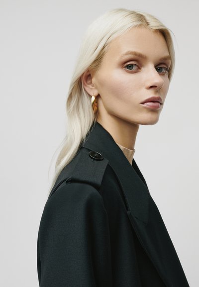 Blonde vrouw met een lichte huid, gekleed in gouden bladeren oorbellen en een zwarte jas, kijkt zijwaarts tegen een effen lichte achtergrond.