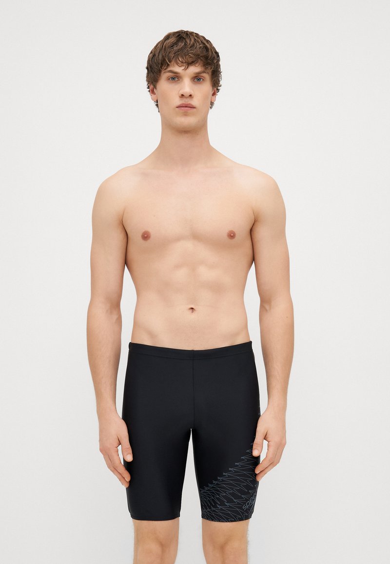 Jovem em pé contra um fundo simples, vestindo calções de banho pretos até aos joelhos com um padrão geométrico subtil numa das pernas.