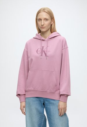 Persoon met steil blond haar draagt een lichtroze Calvin Klein-hoodie en blauwe spijkerbroek, staand tegen een effen lichte achtergrond.