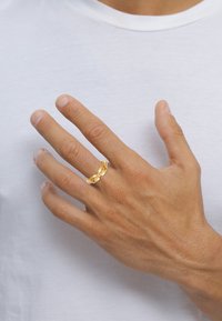 Guldring med sammanlänkad design och klara detaljer. Buren på en hand mot en enkel vit bakgrund. Texturerad hud syns.