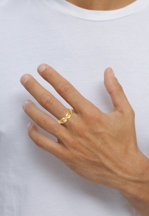 Guldring med sammenflettet design og klare accenter. Båret på en hånd mod en ensfarvet hvid baggrund. Struktureret hud er synlig.