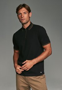 Polo noir avec un col zippé et des accents rayés contrastants sur le col et les manches, associé à un pantalon beige.