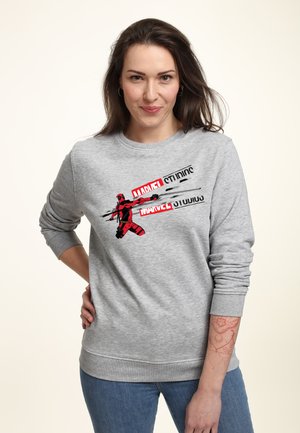 Kobieta o długich brązowych włosach, ubrana w szarą bluzę Marvel Studios z grafiką Deadpoola i niebieskie dżinsy, pozuje z ręką na biodrze.