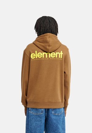 Personne aux cheveux foncés tressés portant un sweat à capuche marron avec un texte jaune "element skateboard co worldwide" dans le dos et un jean bleu.