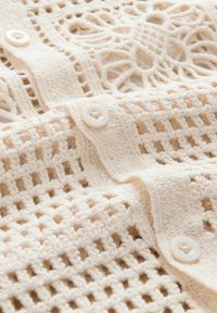 Vêtement tricoté de couleur crème avec un motif en maille ouverte, comportant deux grands boutons et un tissu texturé. Conception légère et aérienne.