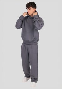 Grå hættetrøje og matchende joggersæt lavet af blødt stof. Hættetrøjen har en forlomme og ribbede manchetter. Der bæres hvide sneakers.