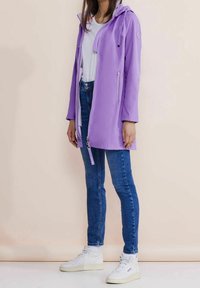 Lila, mjuk skaljacka med huva, dragkedja fram, och sidofickor, som bärs över en vit topp och blå jeans. Vita sneakers fulländar looken.