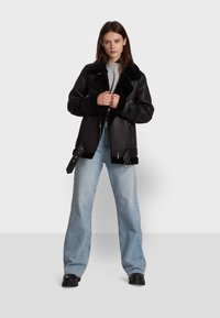 Black faux leather jacket with a shearling collar, zippered pockets, and adjustable straps. Paired with light blue wide-leg jeans.
