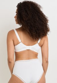 Seno in pizzo bianco con bordo, dotato di spalline regolabili, che mette in mostra un materiale liscio e chiusura a gancio e asola sul retro. Mutandine abbinato a vita alta.