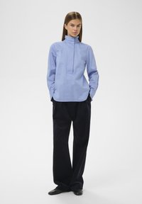 Chemise bleu clair à manches longues avec col montant, patte de boutonnage à l'avant et coupe décontractée, associée à un pantalon large et foncé ainsi qu'à des chaussures noires.