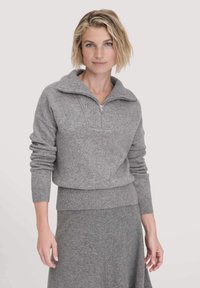 Blonde Frau mit kurzhaariger Frisur, die einen grauen Strickpullover mit Viertelreißverschluss und einen passenden grauen Rock trägt, vor einem schlichten Hintergrund stehend.