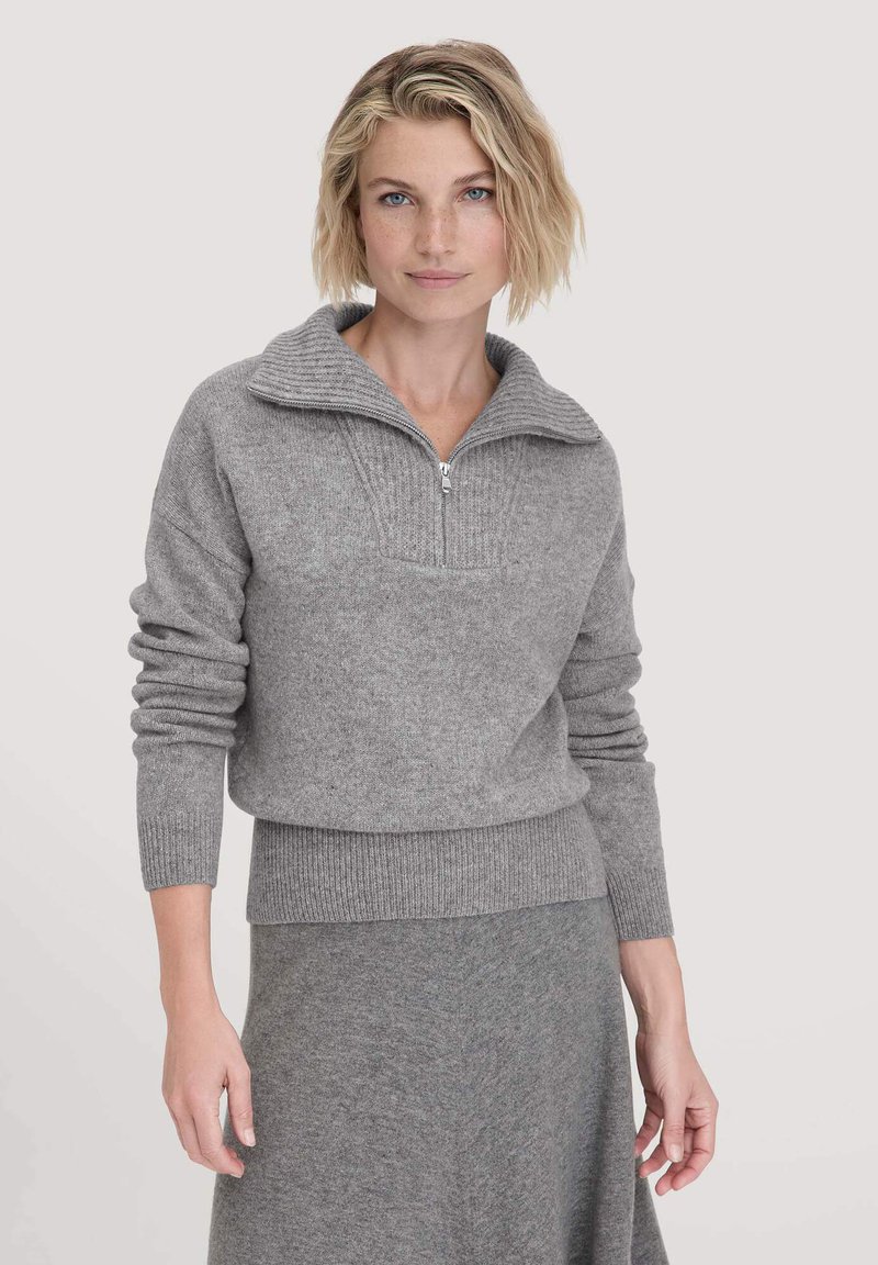 Blonde Frau mit kurzhaariger Frisur, die einen grauen Strickpullover mit Viertelreißverschluss und einen passenden grauen Rock trägt, vor einem schlichten Hintergrund stehend.