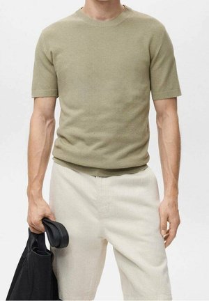 Homme portant une chemise beige texturée à manches courtes et un short de couleur claire, tenant une veste noire dans sa main gauche.