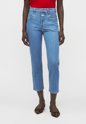 Person trägt hellblaue hoch taillierte kurze Jeans, rotes Oberteil und braune spitze Ballerinas mit Leopardenmuster vor weißem Hintergrund.
