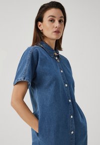 Κοντό τζιν φόρεμα με κοντά μανίκια, κουμπιά μπροστά και πλαϊνές τσέπες. Μπλε ύφασμα με γιακά και πολύχρωμα διακοσμητικά καρφίτσια.