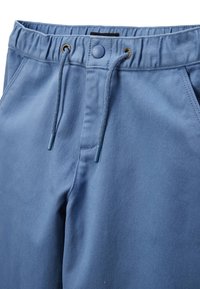 Jogging en mélange de coton bleu avec une taille élastique, un cordon de serrage et des œillets dorés, présentant une fermeture par un bouton unique et des poches latérales.