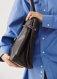 Braune Lederhandtasche mit glänzender Oberfläche, in dreieckiger Form, mit einem silbernen Schnallenakzent und von einer Person mit einem blauen Hemd gehalten.