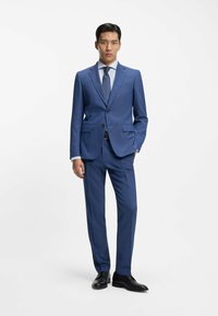 Completo blu con vestibilità slim, giacca monopetto, risvolti a lancia e pantaloni a tinta unita con pieghe piatte. Camicia bianca e cravatta a motivo. Scarpe nere.