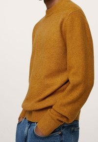 Musztardowy żółty sweter wykonany na drutach z okrągłym dekoltem, prążkowanymi mankietami i dołem, w połączeniu z niebieskimi dżinsami. Widoczna miękka tekstura.