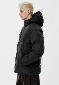 Črno puhovko z visokim ovratnikom in nastavljivo kapuco. Ima gladko, sijočo površino in rahlo oblazinjeno teksturo.