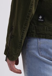 Olivgrüne, leichte Jacke mit einer Reißverschlusstasche am Ärmel und einem Logopatch. Kombiniert mit hellblauen Jeans.